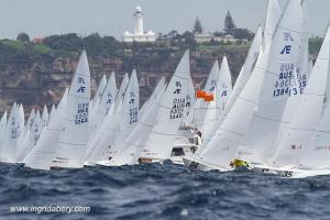 A fleet of white sailboats with visible sail numbers compete in a race on choppy water, with a coastal town and white lighthouse visible in the background.