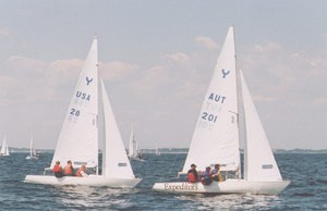 Two small sailboats with white sails, labeled USA 28 and AUT 201, sail side by side on open water. Four people are visible on each boat under a clear blue sky. Other boats appear in the background.