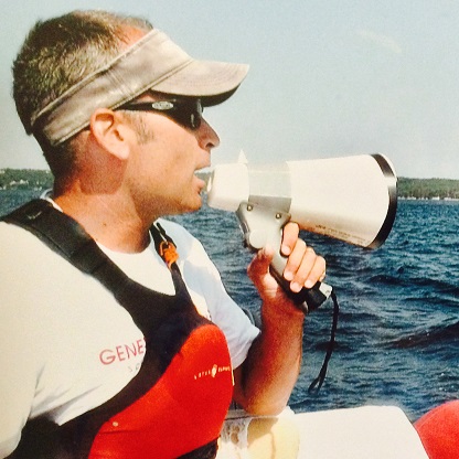 A man wearing sunglasses, a visor, and a life jacket uses a megaphone while sitting in a boat on a body of water.