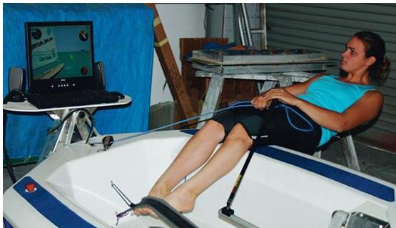 A woman sits in a small boat simulator indoors, holding ropes, with her feet braced, facing a computer screen displaying a sailing simulation.