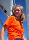 A young person with long hair wearing an orange T-shirt sits on a sailboat, looking off into the distance on a sunny day with a clear blue sky.