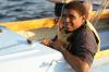 A person wearing a life jacket sits on a small sailboat, smiling at the camera, with water and another yellow boat visible in the background.