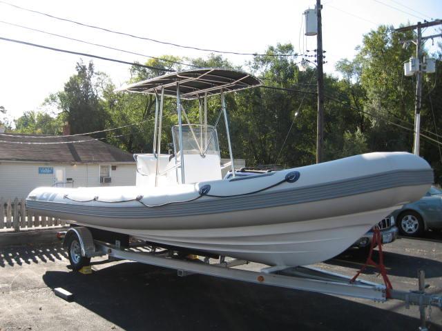 A white inflatable rigid-hull boat with a center console and metal canopy sits on a trailer in a paved lot, with trees, power lines, and a building in the background.