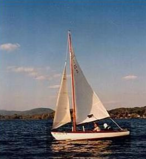 A small sailboat with white sails glides on a calm lake under a blue sky with scattered clouds, with hills and trees in the background.