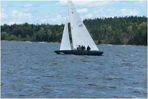A small sailboat with several people on board sails on a lake, with a forested shoreline and a partly cloudy sky in the background.