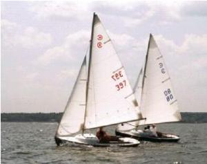 Two small sailboats with white sails glide side by side on a calm body of water under a cloudy sky. Each boat has numbers printed on its sail and carries two people. Trees line the distant shore in the background.
