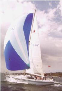 A sailboat with a large blue and white spinnaker sail moves quickly across the water under a cloudy sky, with several people on board.
