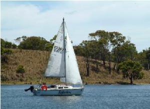 A white sailboat with two sails glides on a calm river, with trees and grassy hills in the background under a partly cloudy sky.