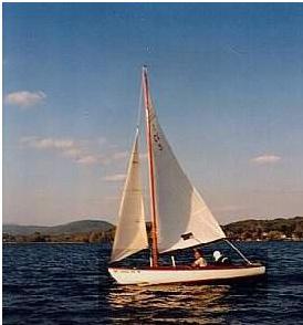 A small sailboat with white sails glides on a calm lake under a blue sky with scattered clouds, with tree-covered hills in the background.