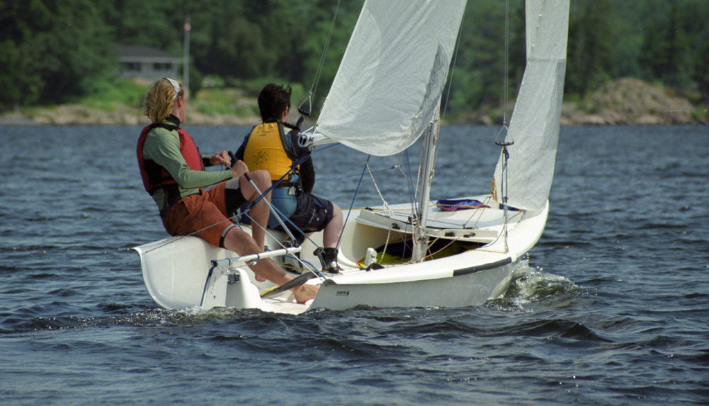 Two people wearing life jackets are sailing a small white sailboat on a lake, surrounded by trees and a house visible in the background.