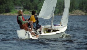 Two people wearing life jackets sail a small white sailboat on a lake, surrounded by trees and greenery in the background.