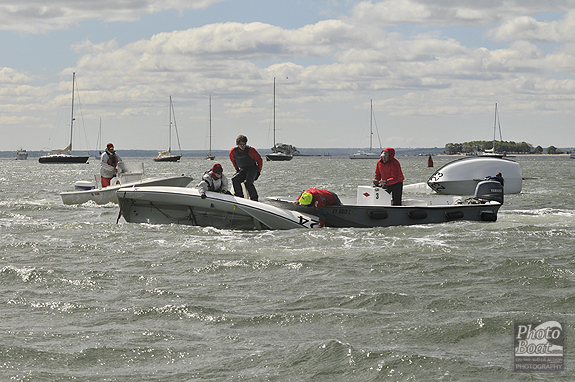 Several people in red jackets assist with a capsized sailboat on choppy water, while other boats and crew are nearby. Sailboats and a distant shoreline are visible in the background under a cloudy sky.