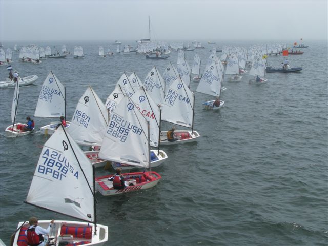 A large group of small sailboats with young sailors participate in a regatta on a calm, open body of water, with many more boats and support vessels visible in the background.