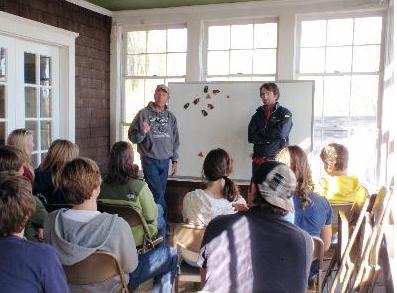 Two people stand in front of a whiteboard, speaking to a seated group in a sunlit room with large windows. The audience appears attentive and engaged.