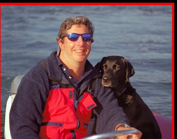 A man wearing sunglasses and a red life jacket smiles while sitting on a boat, with a black dog sitting beside him. The background shows water.