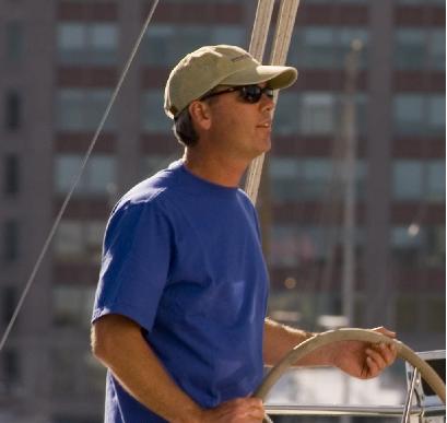 A person wearing a blue shirt, khaki cap, and sunglasses is steering a boat. Blurred buildings are visible in the background.