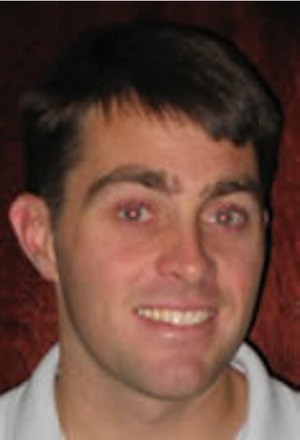 A man with short brown hair and fair skin is smiling at the camera. He is wearing a white collared shirt and is positioned against a dark reddish-brown background.