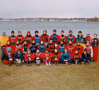 A group of about 30 people in red and blue outdoor gear pose for a photo on grass near a body of water, with buildings and trees visible in the distant background under a cloudy sky.