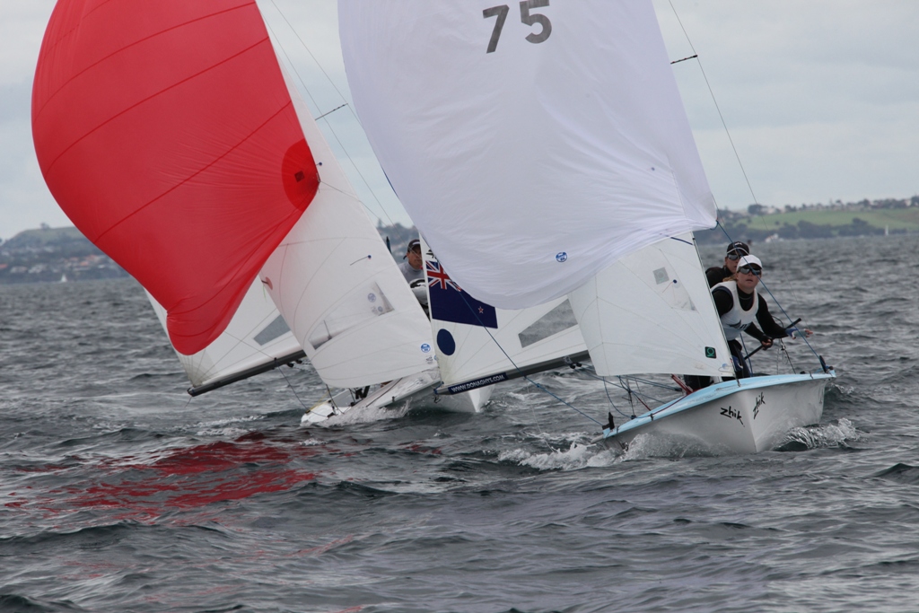 Two small sailboats with spinnakers—one red, one white—race closely on a choppy, gray body of water. Several sailors are visible, and a coastline with houses is in the background beneath a cloudy sky.