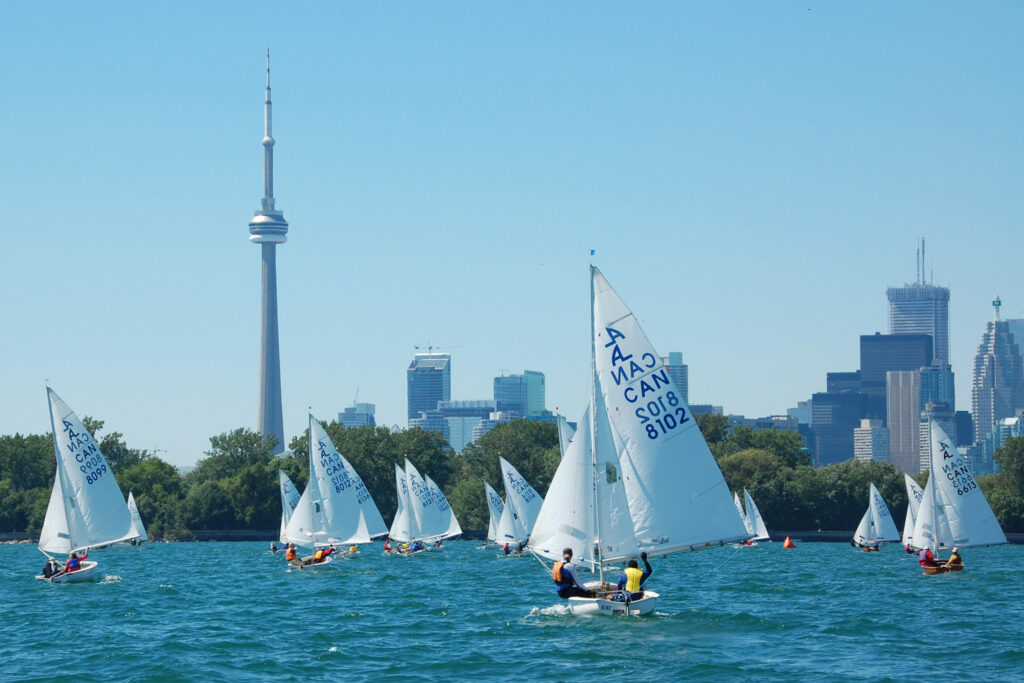 Sailboats with people on a blue lake, racing near the Toronto skyline. The CN Tower and other tall buildings are visible under a clear, sunny sky. Trees line the shore in the background.