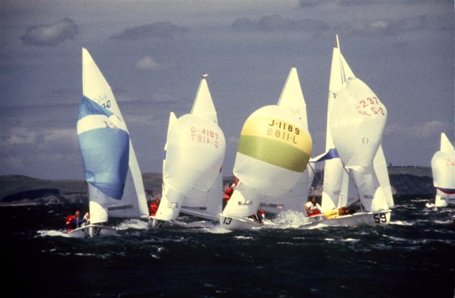Several sailboats with colorful spinnaker sails race closely together on choppy water under a cloudy sky, with distant hills visible in the background.