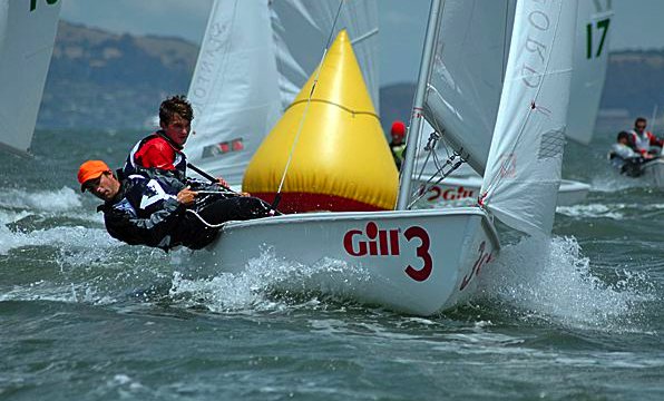 Two sailors are leaning out of a small sailboat labeled 'Gill 3' as it races near a yellow triangular buoy, with other sailboats and choppy water in the background.
