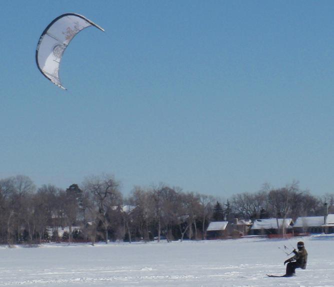A person is snowkiting on a snowy field, holding onto a large kite while gliding on skis. Trees and houses are visible in the background under a clear blue sky.