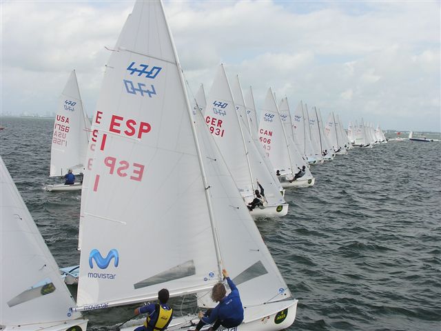 A fleet of sailboats with white sails, each marked with country codes, race closely together on choppy water under a cloudy sky. Sailors are actively adjusting their sails.