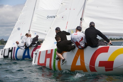 Two sailboats with colorful 'CHICAGO' lettering race side by side on the water, each crew leaning over the edge to balance the boats. The sky is partly cloudy, and land is visible in the background.