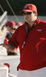 A person wearing a red polo shirt, matching red cap, and sunglasses stands outdoors, appearing to give instructions. A whistle hangs from their neck, and other people in red outfits are visible in the background.