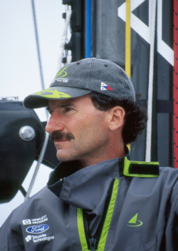A man with a mustache wearing a gray cap and a gray sailing jacket stands on a boat, looking to the left. The jacket and cap have green accents, and there are sponsor logos on his sleeve. Boat equipment is visible in the background.