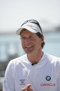 A man in a white ORACLE and BMW-branded shirt and white cap stands outdoors, smiling. He is wearing sunglasses on his cap, with a blurred waterfront in the background.