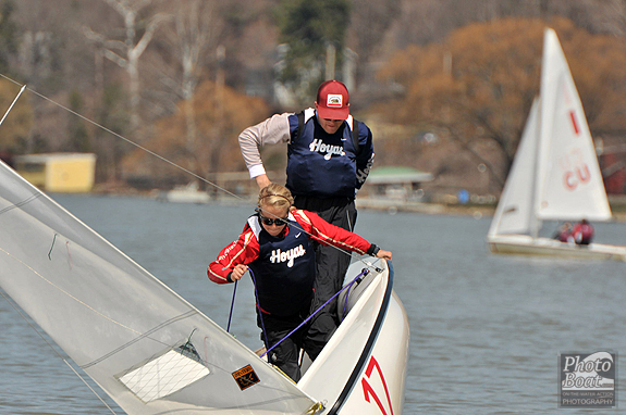 Two people in 'Hoyas' jerseys are sailing a boat, with one standing at the bow and the other behind, both focused. Other sailboats and a shoreline with trees are visible in the background.