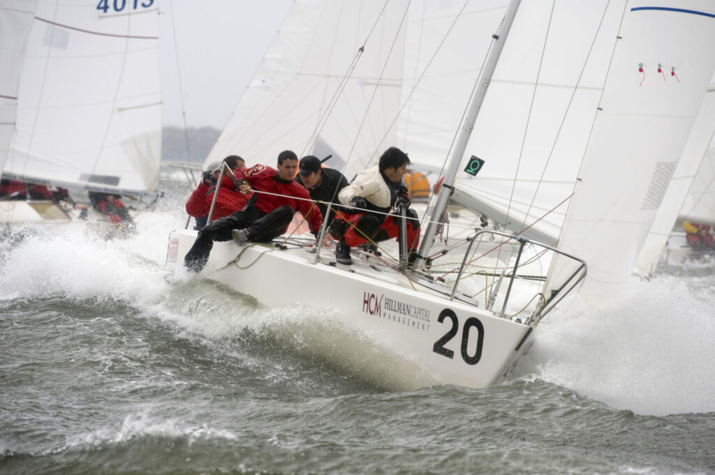 Four sailors in wet-weather gear lean over the side of a white sailboat numbered 20, racing through choppy waves with water spraying up as other sailboats compete in the background.