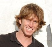 A smiling man with medium-length, tousled light brown hair wears a dark collared shirt and stands in front of a light-colored background.