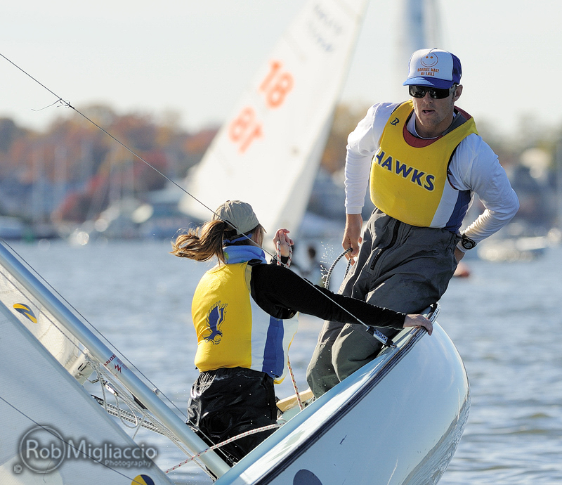 Two sailors in yellow vests, marked 'HAWKS,' maneuver a sailboat on a sunny day. The man stands on the edge, leaning out, while a woman adjusts the ropes. Other sailboats and blurred autumn trees are in the background.