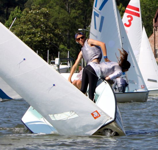 Two sailors lean out of a small sailboat sharply tilted on the water, competing in a regatta. Other sailboats and trees are visible in the background. The scene suggests action and balance in sailing.