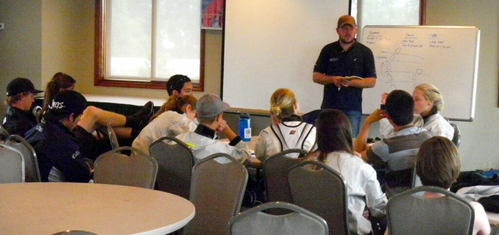A group of people sit around tables in a classroom, listening to a person standing in front of a whiteboard with diagrams and notes. Some attendees have notebooks and water bottles.