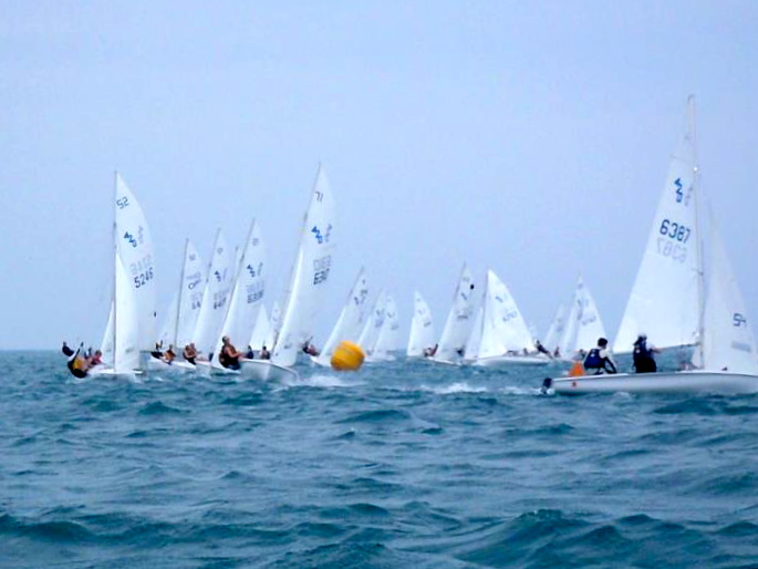 Several small sailboats race across choppy blue water, each with white sails marked by numbers. Sailors lean out to balance their boats, and a yellow buoy floats among them under a cloudy sky.