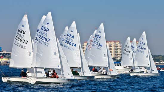 Several sailboats with visible country codes (URU, ESP, USA) compete in a race on the water, with multiple sailors aboard each boat. Buildings and a clear blue sky are in the background.