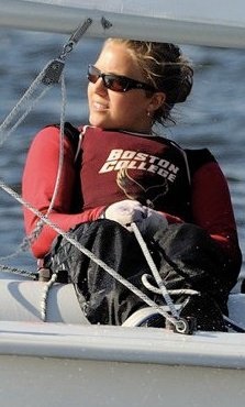 A woman wearing sunglasses and a 'Boston College' shirt is sailing a boat, holding ropes and leaning back, with water in the background.