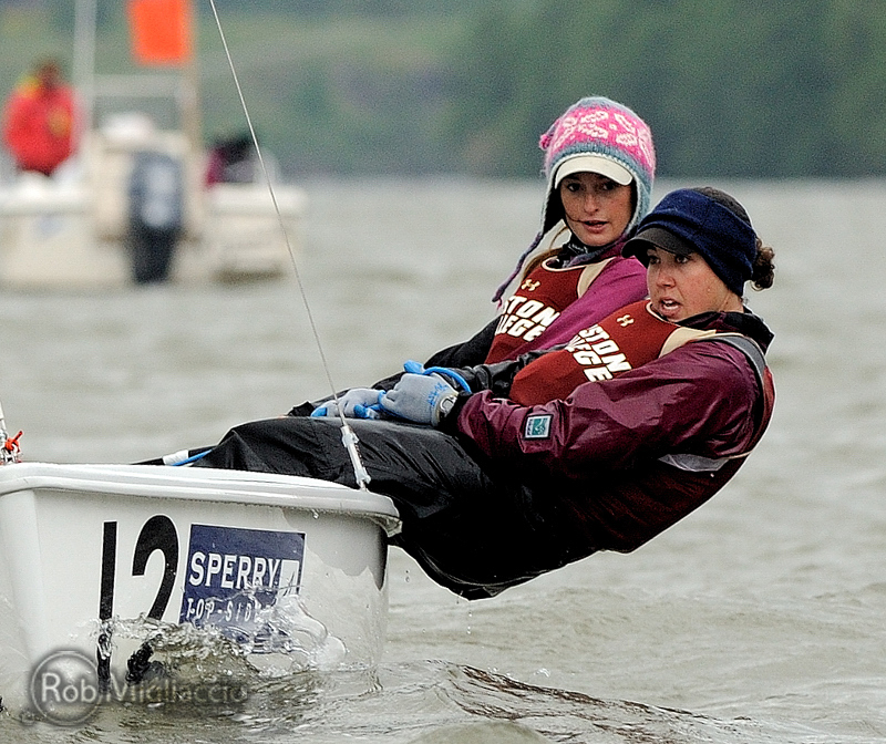 Two women in maroon Boston College sailing jackets balance on the edge of a small sailboat, leaning out over the water during a race. Both wear hats and life vests, and another boat is visible in the background.
