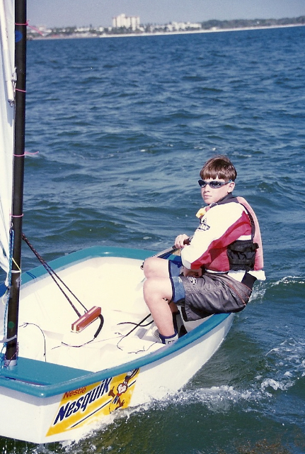 A boy wearing sunglasses and a life jacket sails a small boat with a Nesquik logo on the side, gliding over blue water on a sunny day. A distant shoreline is visible in the background.