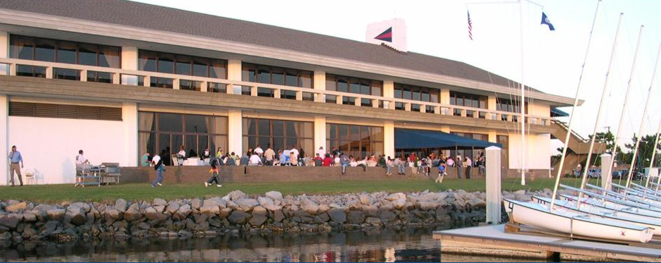 A waterfront building with large windows and a stone shoreline, people gathered outside on the grass, and several sailboats docked in the foreground.