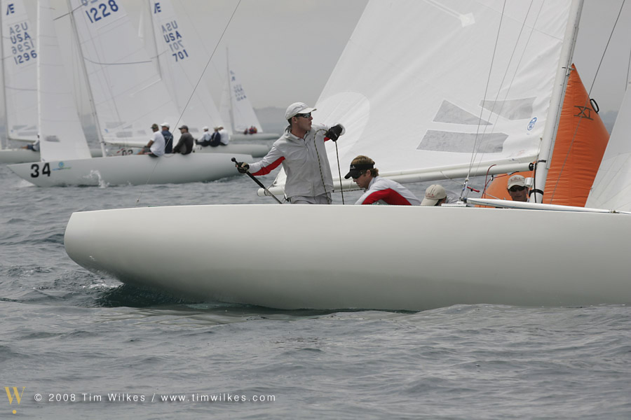Several sailors compete in a regatta, maneuvering a white sailboat with orange and white sails on a gray, overcast day. Other sailboats can be seen in the distance on the water.