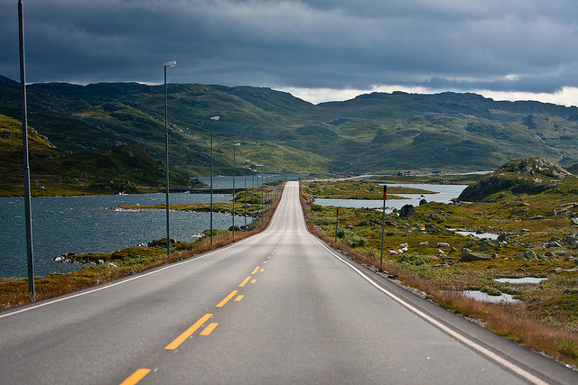 A long, empty road stretches into the distance between lakes and green hills under a cloudy sky, with streetlights lining the roadside.