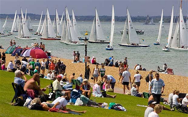 A crowded beach scene with people relaxing on grass and pebbles while many sailboats race on the water in the background under a sunny sky.
