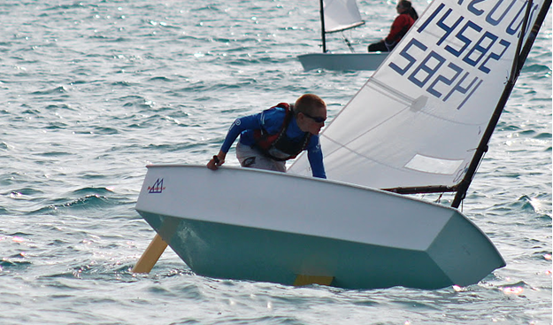 A young sailor leans out of a small white sailboat on choppy blue water, balancing the boat as it tilts. Another sailor is visible in the background, also navigating a similar boat.