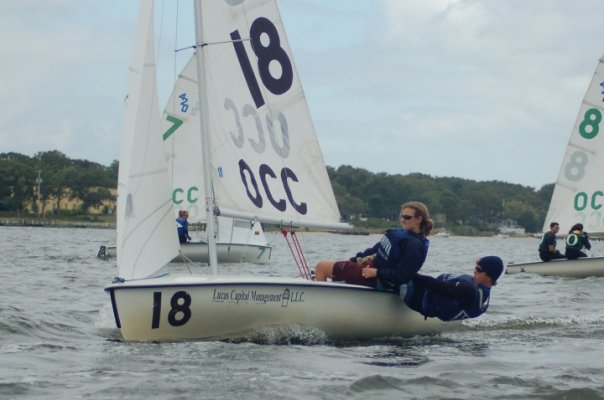 Two people sail a small boat labeled '18' and 'OCC' on a lake, leaning back for balance. Other sailboats are visible on the water, with trees and buildings in the background under a cloudy sky.