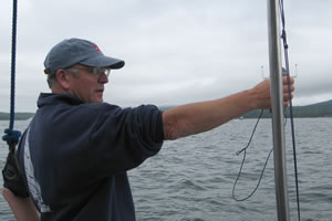 A person wearing a cap and glasses stands on a boat, holding a device with wires over the water, with a cloudy sky and distant shoreline in the background.